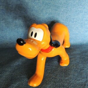 May include: A ceramic figurine of Pluto, a cartoon dog from Disney. Pluto is orange with black ears and a red collar. He is standing on all fours with his head tilted to the side.