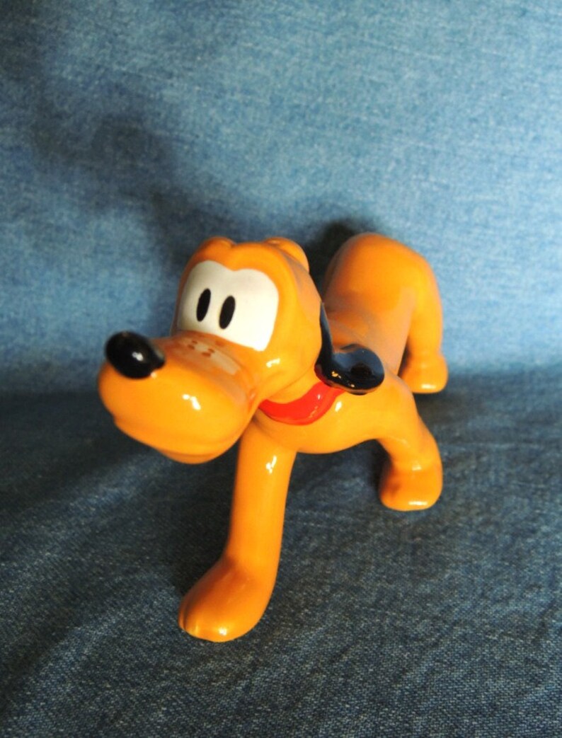 May include: A ceramic figurine of Pluto, a cartoon dog from Disney. Pluto is orange with black ears and a red collar. He is standing on all fours with his head tilted to the side.