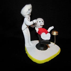 May include: A whimsical figurine depicts two skeleton figures in a barber shop scene. One skeleton, dressed in white, is cutting the hair of another skeleton seated in a black chair, wearing a red shirt. The base is white with a yellow border.