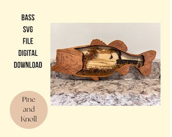 May include: A wooden bass fish cutout with a scene of a fisherman in a boat inside the fish. The scene is made of wood and has a sunset background.