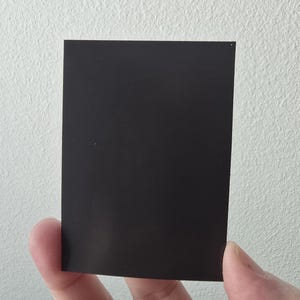 Puede incluir: Un im&aacute;n rectangular negro liso, sostenido contra una pared blanca texturizada. El im&aacute;n tiene una forma simple y plana, con un acabado mate. La imagen es un primer plano, que destaca el color y la textura del im&aacute;n.
