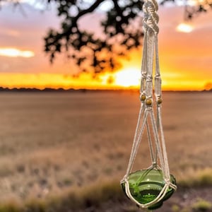 May include: A macrame plant hanger crafted from off-white cord, featuring intricate knotted patterns and small wooden beads. The hanger supports a small, translucent green glass bowl with a scalloped rim, suspended by a metal ring at the top.
