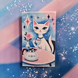 May include: A rectangular illustration of a white cat with blue eyes, wearing a pearl necklace, holding a teacup. The cat sits at a pink table with a coffee pot and a vase of pink flowers. The background is blue with starburst and star accents.