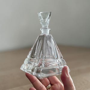 May include: A clear, faceted glass perfume bottle with a pyramid-shaped body and a triangular stopper. The bottle is held in a hand, showcasing its intricate design and vintage aesthetic.