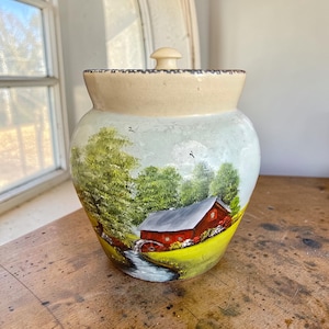 May include: A hand-painted ceramic jar with a lid. The jar features a landscape scene with a red barn, green trees, and a flowing stream. The top and lid are a cream color, and the jar sits on a wooden surface.