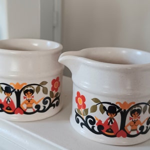 May include: A set of two white ceramic creamer and sugar containers with a folk art design featuring stylized figures in red, orange, yellow, green, and black.