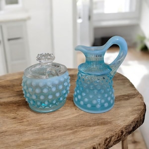 May include: A light blue hobnail glass sugar bowl with a lid and a small creamer pitcher. The sugar bowl and creamer are made of glass and have a hobnail pattern. The sugar bowl has a lid with a decorative knob.