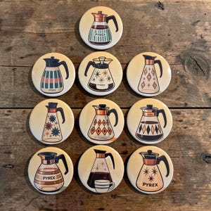 May include: A collection of nine round Pyrex-themed buttons with various designs. Each button features a different illustration of a Pyrex coffee pot in shades of brown, black, and cream, with the word "PYREX" printed on some.