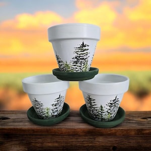 May include: Three white ceramic flower pots with a hand-painted forest design in black and green. Each pot sits on a dark green saucer. The pots are arranged on a wooden surface with a blurred sunset background.