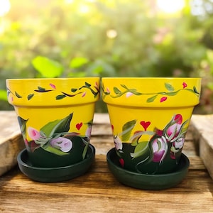 May include: Two hand-painted yellow flower pots with matching dark green saucers. The pots feature a floral design with pink and white flowers, green leaves, and red heart accents. The pots are sitting on a wooden surface.