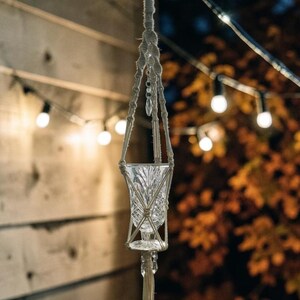 May include: A clear glass votive holder with a diamond-cut pattern suspended in a beige macrame hanger. The hanger features intricate knotted cord work and decorative clear crystal-like beads integrated into the vertical support strands.