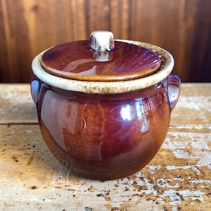 May include: A small, round, brown ceramic pot with a lid. The pot has a glossy finish with a lighter-colored rim and a silver-colored knob on the lid. Two small handles are on the sides of the pot.