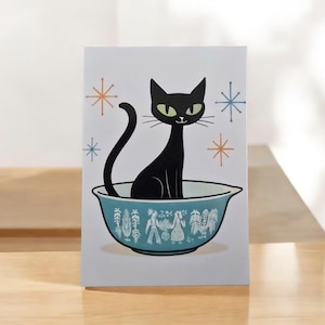 May include: A greeting card featuring a black cat sitting in a teal bowl with a decorative rim. The cat has green eyes and is surrounded by starburst designs in orange and blue. The card has a white background.