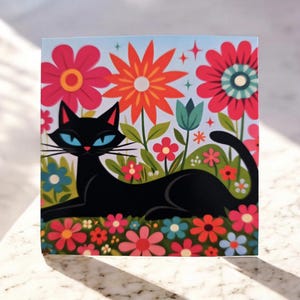 May include: A square art print featuring a stylized black cat with blue eyes, reclining in a colorful garden of flowers. The flowers are in shades of red, orange, pink, and blue, set against a light blue background.