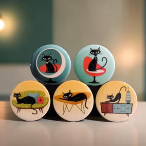 May include: Five circular buttons featuring illustrations of black cats in various settings. The top two show cats on chairs against blue and teal backgrounds. The bottom three show cats on a green sofa, a yellow table, and a blue cabinet.