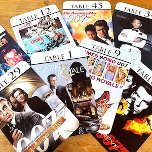 May include: A collection of James Bond movie-themed table cards. Each card features a different movie poster, with titles like "Casino Royale" and "Goldfinger." The cards are numbered, with titles like "Table 12" and "Table 45."
