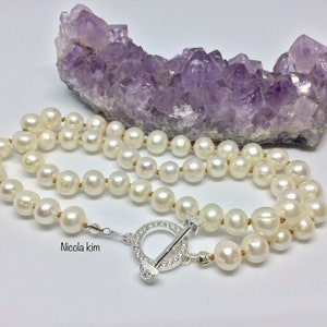 May include: A white pearl necklace with a silver toggle clasp. The necklace is displayed on a purple amethyst crystal.
