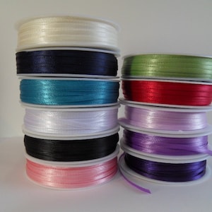 May include: A stack of spools of satin ribbon in various colours, including white, black, teal, light pink, lavender, and purple.