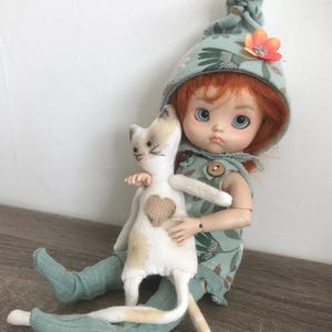 May include: A doll with red hair and blue eyes wearing a green outfit and hat, holding a white cat plushie with a heart. The doll's outfit has a floral pattern and a flower on the hat.