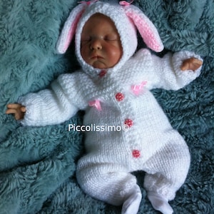 May include: A white knitted bunny rabbit outfit with pink ears and pink bows. The outfit has red buttons with white polka dots. The outfit is on a light blue fluffy blanket.
