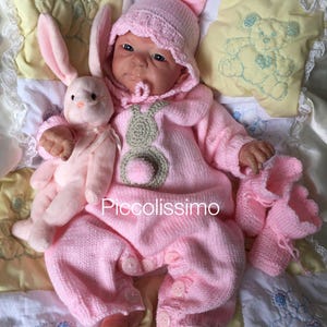 Può includere: Un completo per neonato in maglia rosa con cappello e scarpine abbinati. Il completo presenta un design a coniglietto e un pon pon sul davanti. Il bambino tiene un peluche rosa a forma di coniglietto. Il testo "Piccolissimo" è visibile sul completo.