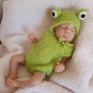May include: A lifelike baby doll wearing a lime green knitted frog costume with a hood and large, round eyes. The doll is lying on a soft, white blanket. The costume covers the baby's body and has a small tie at the neck.