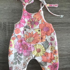 Könnte beinhalten: Ein Strampler mit Blumenmuster in Weiß, Rosa, Orange und Gelb. Der Strampler hat eine Tasche und Bindeträger. Der Stoff ist mit großen Blumen und kleineren floralen Akzenten bedeckt.