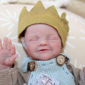 May include: A lifelike baby doll wearing a yellow crown and a blue and brown outfit. The doll has closed eyes and a slight smile. The outfit has wooden button details. The doll is lying on a patterned beige surface.