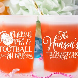 May include: Two orange plastic cups with white text. One cup says "Turkey, Pie & Football - Oh My!" with a turkey and football graphic. The other cup says "The Hanson's Thanksgiving 2018" with a leaf design.