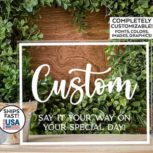 May include: A clear acrylic sign with a white border and the word "Custom" written in a cursive font. The text "Say it your way on your special day!" is written below the word "Custom".