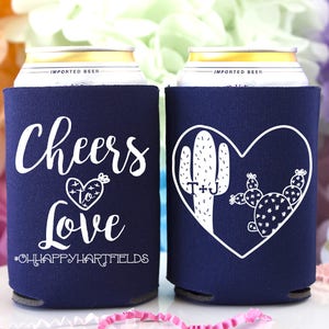 May include: Two navy blue can coolers with white text and designs. One cooler says "Cheers to Love #OHHAPPYHARTFIELDS". The other cooler has a heart with a cactus and the text "T+J".