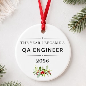 May include: A round white ceramic ornament featuring a red ribbon for hanging. The text reads THE YEAR I BECAME A QA ENGINEER 2026 in black lettering, accented by a small illustration of green leaves and red berries at the bottom of the ornament.