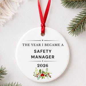 May include: A round white ceramic ornament featuring a red ribbon for hanging. The text reads THE YEAR I BECAME A SAFETY MANAGER 2026, accented with a small illustration of green holly leaves and red berries at the bottom of the ornament.