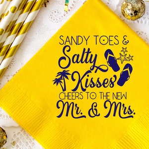 May include: Yellow paper napkin with the text "Sandy Toes & Salty Kisses" in blue lettering. Below the text is a pair of blue flip flops and two palm trees. The text "Cheers to the new Mr. & Mrs." is below the flip flops.