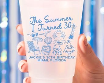 May include: A frosted plastic cup with blue text and illustrations. The text reads "The Summer I Turned 30" with beach-themed graphics. Below, it says "JACKIE'S 30TH BIRTHDAY, MIAMI, FLORIDA, Est. 1996". The cup is held in a hand.