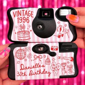May include: Two disposable cameras with black plastic bodies and custom light pink and white striped wraps. The top camera features red text reading VINTAGE 1996 and a birthday cake. The bottom camera reads Danielle's 30th Birthday with disco ball graphics.