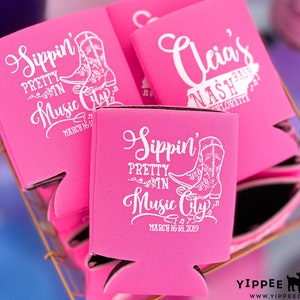 May include: Pink can coolers with white text and a boot design. The text reads "Sippin' Pretty in Music City" and "March 16-18, 2019".