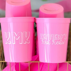 May include: Two pink plastic cups with white lettering. One cup says "KMK" and the other says "Kennedy's First Fiesta!"