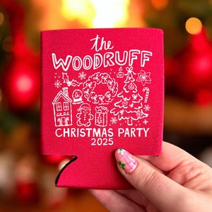 May include: A red foam can cooler featuring white line art illustrations of a house, a gingerbread person, a wreath, and a Christmas tree, with the text "the WOODRUFF CHRISTMAS PARTY 2025" printed in a white hand-lettered style font on the front.