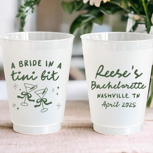May include: Two white plastic cups with green text. One cup says "A bride in a tini bit" with a graphic of two martini glasses. The other cup says "Reese's Bachelorette Nashville, TN April 2025".