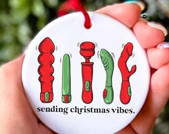 Puede incluir: Adorno navideño de cerámica blanca con un cordón rojo. El adorno presenta una línea de cinco juguetes de dibujos animados coloridos con el texto "sending christmas vibes."