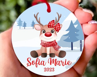 Pode incluir: Enfeite de Natal redondo com uma rena de desenho animado com o nariz vermelho, um laço vermelho com bolinhas brancas e uma echarpe vermelha e branca. O enfeite tem um fundo azul com neve branca e duas pequenas árvores verdes. O texto "Sofia Marie 2023" está impresso no enfeite.