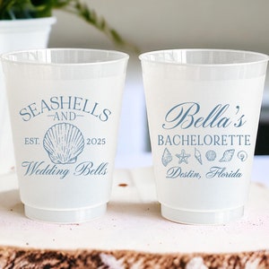 May include: Two white plastic cups with blue text and graphics. The left cup says "Seashells and Wedding Bells" with a seashell graphic and the text "Est. 2025". The right cup says "Bella's Bachelorette" with a starfish and seashells graphic and the text "Destin, Florida".