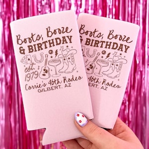 May include: Two pink can coolers with the text "Boots, Booze & Birthday" and a graphic of cowboy boots, a guitar, and a horseshoe. The text "Corrie's 46th Rodeo" and "Gilbert, AZ" are also printed on the coolers.