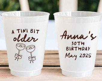 May include: Two white plastic cups with brown text. One cup says "A TINI BIT older" with two martini glasses. The other cup says "Anna's 30TH BIRTHDAY May 2025".