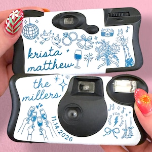 May include: Two black disposable cameras with white custom labels. The top label features blue line art of a disco ball, doves, rings, bells, a bouquet, and champagne, with the text "krista + matthew". The bottom label shows "the millers", "11.14.2026", champagne glasses, and musical notes.
