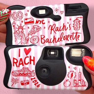 May include: Two disposable cameras with a pink and white striped background. The top camera features red illustrations of a pretzel, pizza slice, donut, Statue of Liberty, and the text "NYC" and "Rach's Bachelorette". The bottom camera shows "I heart RACH", a taxi, "2026", and a city skyline.