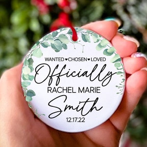 Op de afbeelding: Ronde witte ornament met groene aquarelbladeren en de tekst "WANTED CHOSEN LOVED Officially Rachel Marie Smith 12.17.22"