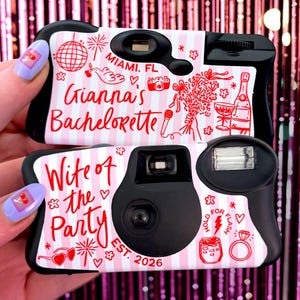 May include: Two black disposable cameras with pink and white striped designs. One camera reads "Gianna's Bachelorette" and the other reads "Wife of the Party". Both cameras have red illustrations and text, including "Miami, FL" and "EST. 2026".