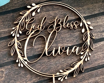 May include: Wooden cake topper with a circular wreath design. The wreath is made of leaves and berries. The text "God Bless Olivia" is written in cursive inside the wreath.